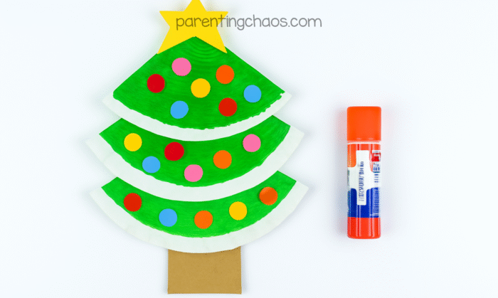 Decorated paper plate Christmas tree craft for kids with glued paper ornaments and a yellow star on top, glue stick shown beside it.