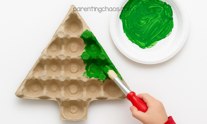 Child painting an egg carton Christmas tree green with a red-handled paintbrush, using green paint on a paper plate.