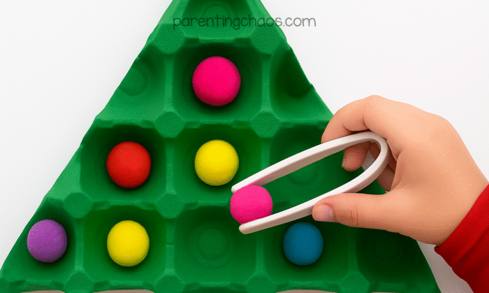 Child using white tweezers to place a pink pom pom ornament on a green egg carton Christmas tree.