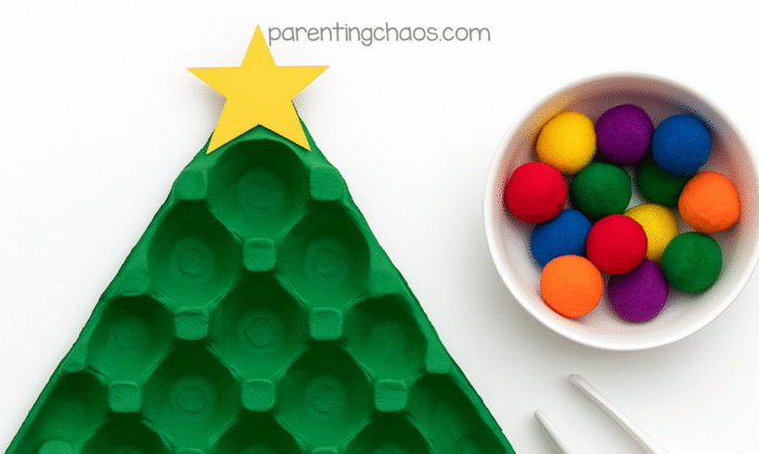 Painted egg carton Christmas tree with a yellow star and a bowl of colorful pom poms and tweezers beside it.