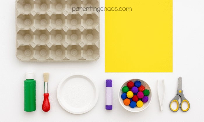 Supplies for Christmas tree pom pom activity, including egg carton, yellow paper, green paint, red-handled brush, glue stick, scissors, tweezers, paper plate, and colorful pom poms.