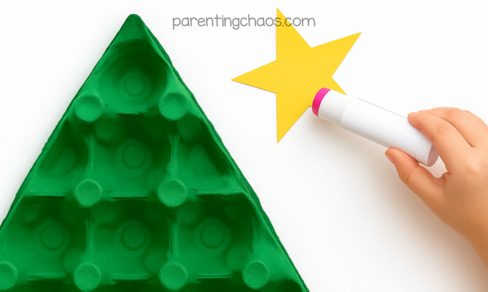 Child using a glue stick to add adhesive to a yellow paper star next to a painted green egg carton Christmas tree.