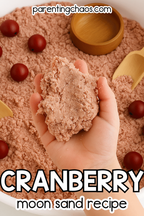 A child’s hand holding a handful of pink cranberry moon sand over a sensory bin filled with red wooden balls and a small wooden scoop. Text reads “Cranberry Moon Sand Recipe.”