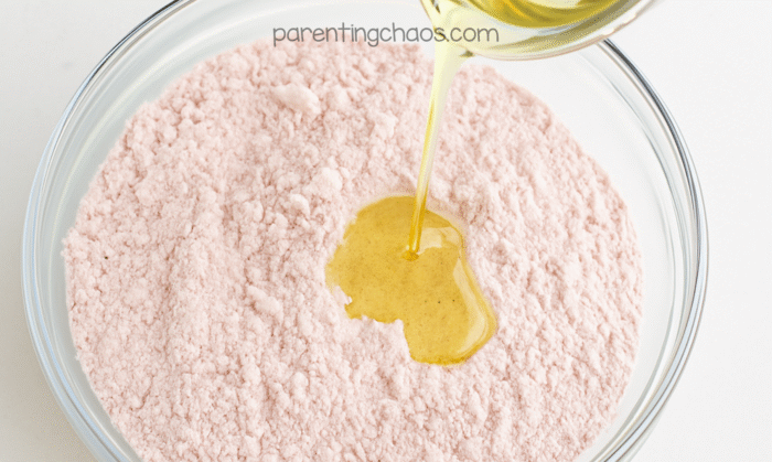 Oil being poured into a bowl of pink cranberry moon sand mixture during the wet ingredient step.
