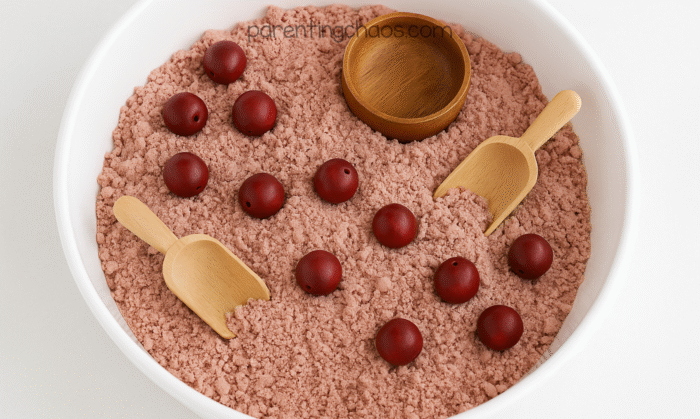 Finished cranberry moon sand displayed in a white sensory bin with red wooden beads, wooden scoops, and a small wooden bowl.