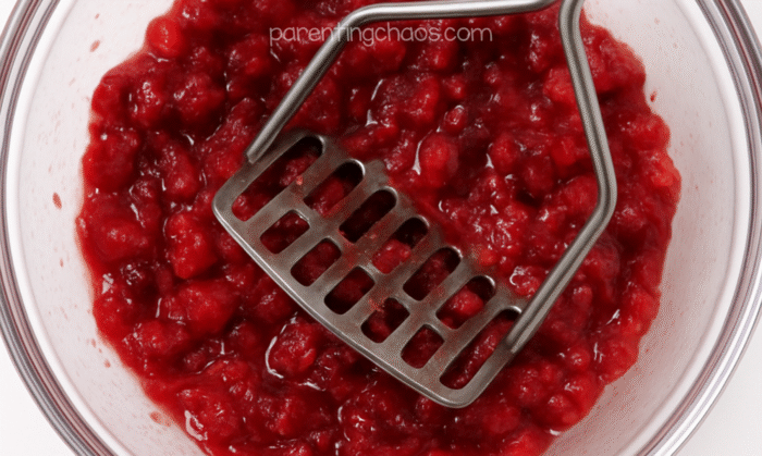 Bowl of mashed cranberries with a metal masher pressing into the mixture.