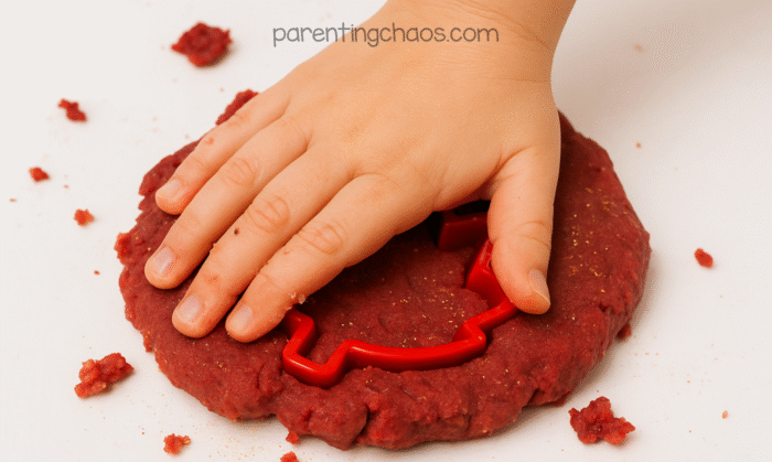 Child’s hand pressing a cookie cutter into red cranberry playdough on a white surface.