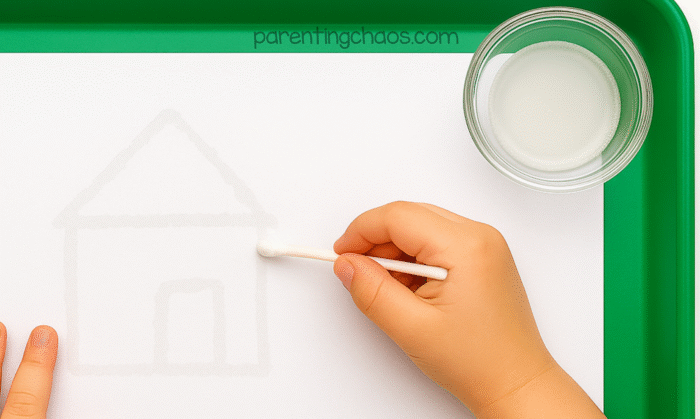 Child using a Q-tip dipped in baking soda solution to draw a house on white paper.