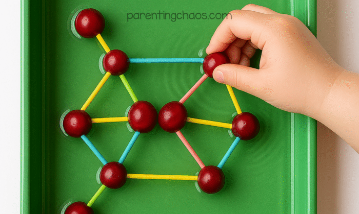 Overhead photo of a child’s hand building a geometric cranberry structure in a green tray filled with water using red cranberries and colorful toothpicks.