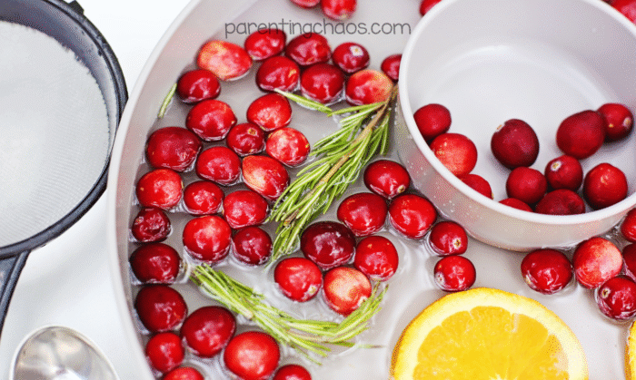 Overhead photo of a cranberry water sensory bin filled with floating cranberries, rosemary sprigs, and orange slices in clear water. A small white bowl and strainer sit in the tray, ready for scooping play.