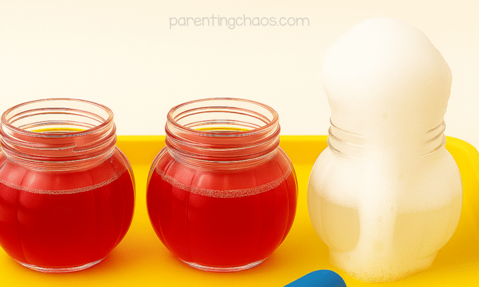 Three jars lined up on a yellow tray — two filled with cranberry juice and one with vinegar — showing the start of the chemical reaction as one begins to foam.