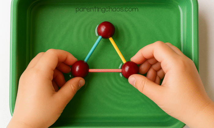 Overhead view of a child’s hands building a triangle with cranberries and colorful toothpicks inside a green sensory tray filled with water.