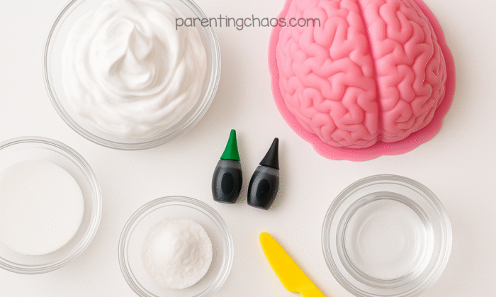 Overhead view of ingredients for fluffy brain slime sensory play, including shaving cream, glue, baking soda, water, black and green food coloring, a yellow plastic knife, and a pink brain mold.