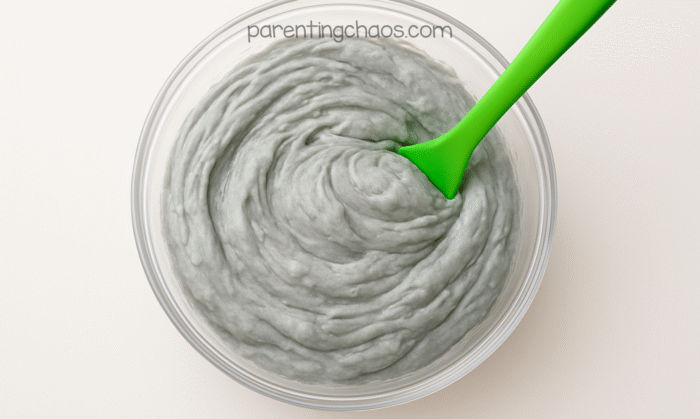 Fluffy gray slime being stirred in a clear bowl with a bright green silicone spoon.