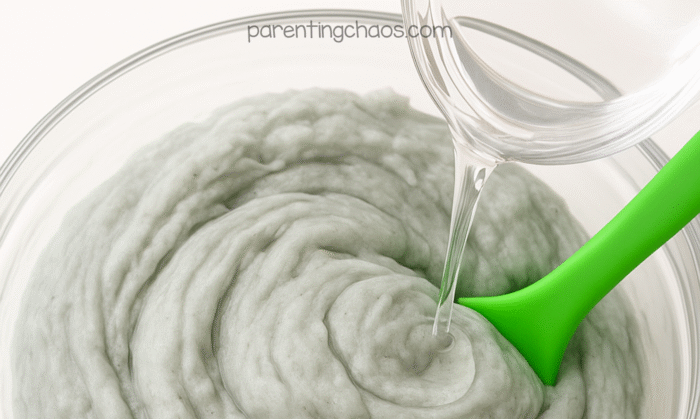 Close-up of contact solution being poured into a bowl of gray fluffy slime with a green spoon for mixing.