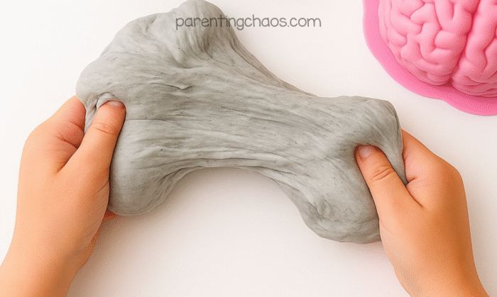 Child’s hands stretching gray fluffy brain slime on a white surface with the pink brain mold nearby.