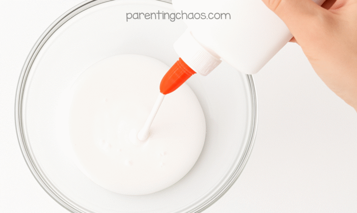 Close-up of white school glue being squeezed into a clear mixing bowl from an orange-tipped bottle.