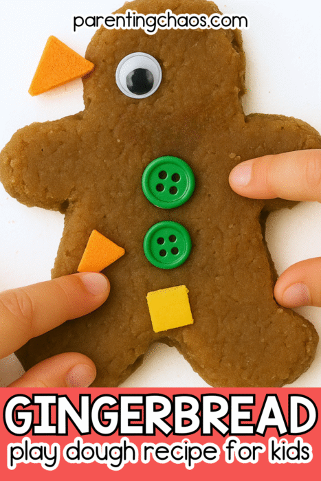 A gingerbread man made from brown play dough. The figure has one googly eye, two green buttons, and orange and yellow foam shapes for decoration. Two small fingers are pressing on the play dough. Text reads “Gingerbread Play Dough Recipe for Kids.”