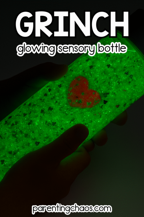Child’s hands holding a green glowing Grinch sensory bottle in the dark, with a red heart charm visible inside.