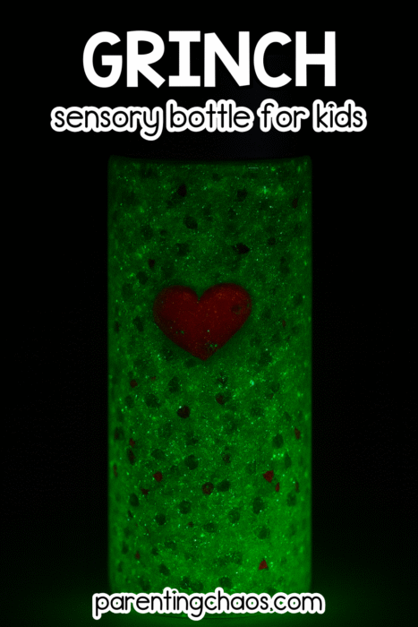 A glowing green Grinch sensory bottle standing in the dark with a bright red heart charm centered inside.
