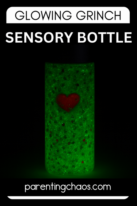 A glowing green Grinch sensory bottle standing in the dark, filled with green glitter and a red heart charm.