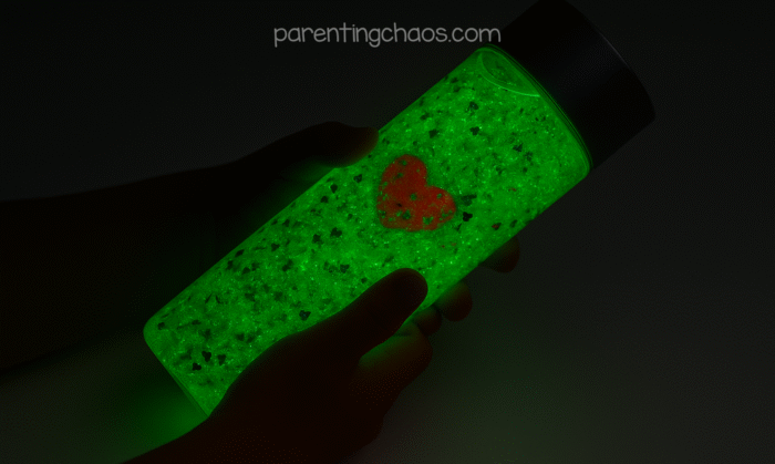 A child holding the green glitter sensory bottle in a dark room, the liquid glowing bright neon green with the red heart charm still visible inside.