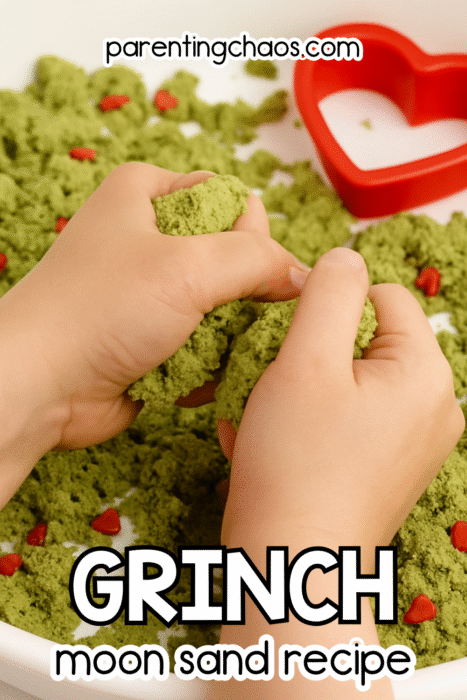 A child squeezing bright green Grinch moon sand in a sensory tray filled with red heart sprinkles and a red heart cookie cutter.