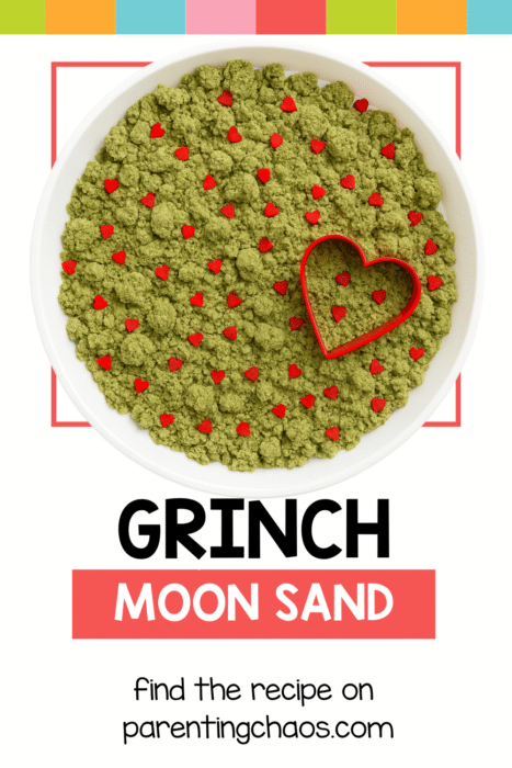 A white tray filled with green Grinch moon sand and red heart sprinkles, with a red heart cookie cutter resting on top.