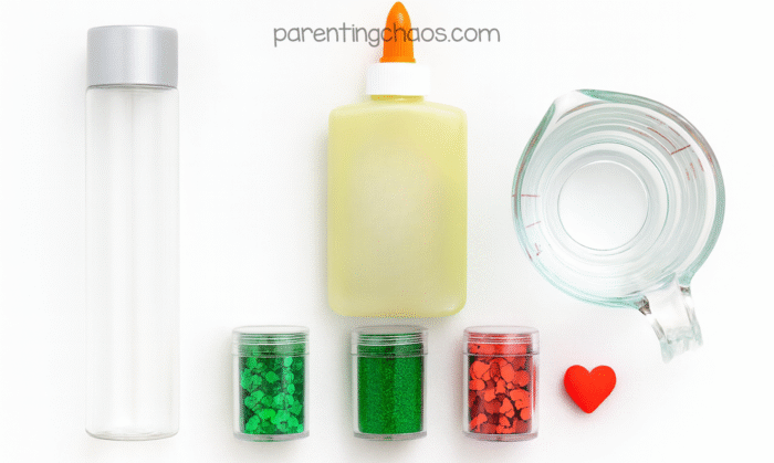 Clear plastic sensory bottle, a bottle of glow in the dark school glue, a glass measuring cup filled with water, three small jars of red and green glitter, and a small red heart charm arranged on a bright white background.