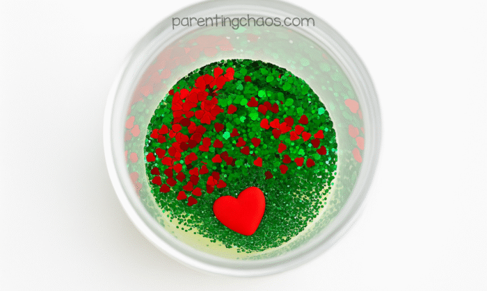 Top-down view into the sensory bottle showing a pile of red and green heart-shaped glitter sitting on top of the glue with a small red heart charm resting in the center.