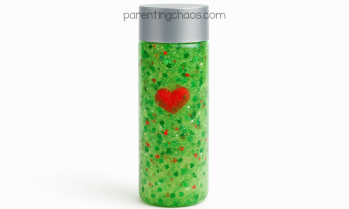 A tall clear sensory bottle filled with bright green Grinch-themed glitter liquid, red and green heart confetti, and a red heart charm floating near the front.