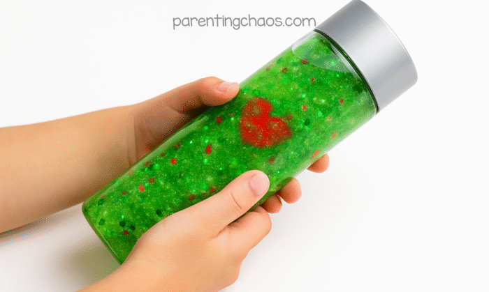 A child’s hands holding a green glitter sensory bottle, with the red heart charm visible through the front of the bottle.