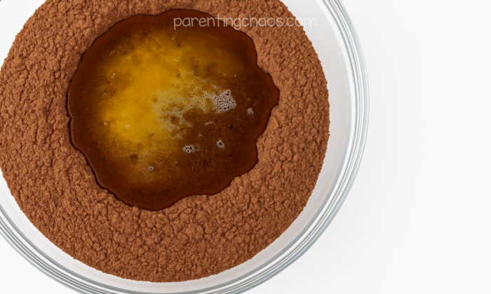 Wet ingredients poured into the center of the cocoa powder mixture in a glass bowl, showing melted oil and vanilla pooling in the middle.