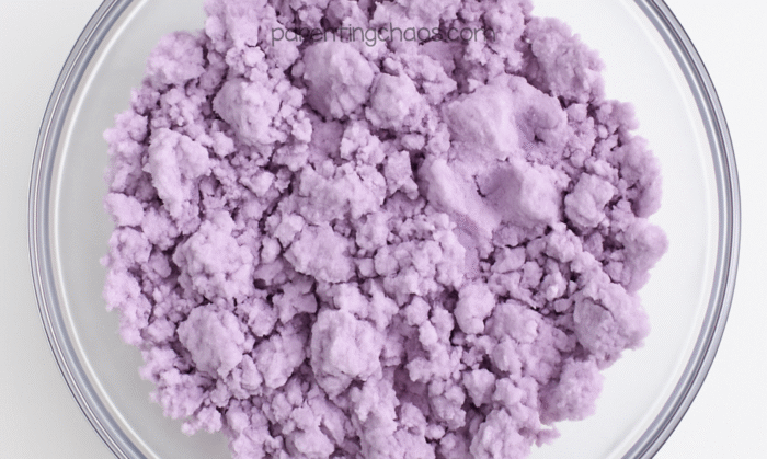 Lavender playdough mixture starting to come together, showing a crumbly purple texture in a clear bowl.