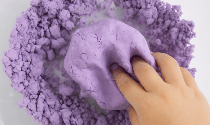 Child’s hand pressing crumbly lavender playdough mixture together as it starts to form into a dough.