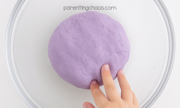 Smooth lavender playdough ball resting in a glass bowl with a child’s finger gently pressing the surface.