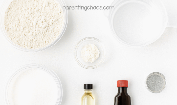 Overhead view of peppermint playdough ingredients, including flour, cream of tartar, salt, oil, peppermint extract, glitter, and water arranged on a white background.