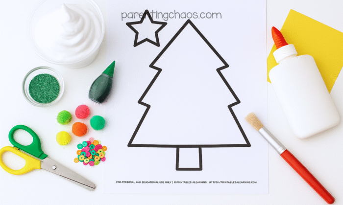 Flat lay of Christmas tree craft supplies including a shaving cream container, green glitter, green food dye, colorful pom poms, sequins, scissors, glue, a paintbrush, and a printable Christmas tree outline on white paper.