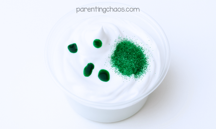 Close-up of a bowl of white shaving cream with drops of green food coloring and a pile of green glitter on top, ready to mix for puffy paint.