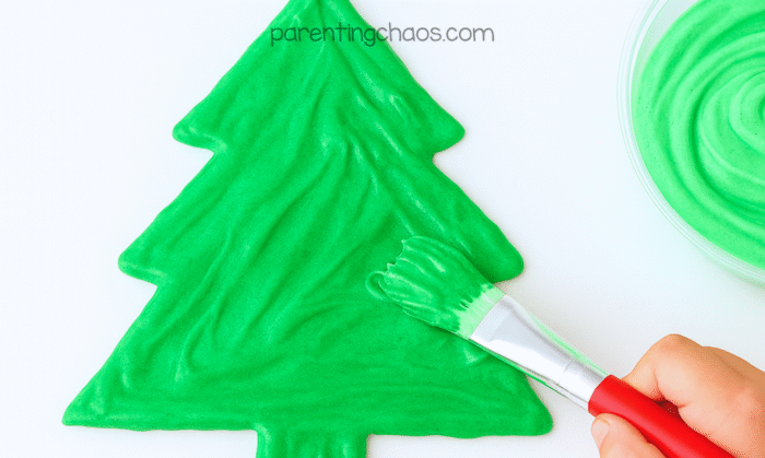 Child’s hand painting the printable Christmas tree shape with green puffy paint using a red-handled paintbrush.