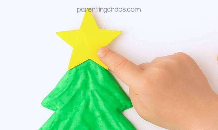 Child’s hand pressing a yellow paper star onto the top of the green puffy paint Christmas tree craft.