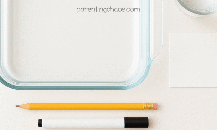 Overhead photo of materials for the floating spiders science experiment — a clear glass dish, pencil, dry erase marker, small piece of white paper, and water cup on a white background.