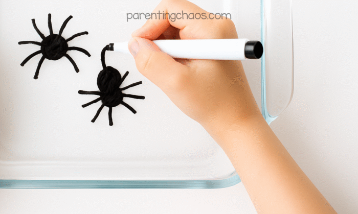 Child drawing two black spiders with a dry erase marker inside a clear glass dish for the floating spiders science experiment.