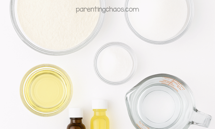 Flat lay of sugar cookie playdough ingredients. Glass bowls hold flour, salt, cream of tartar, oil, and water. Two bottles of extract sit beside the bowls on a bright white background. Text reads “parentingchaos.com.”