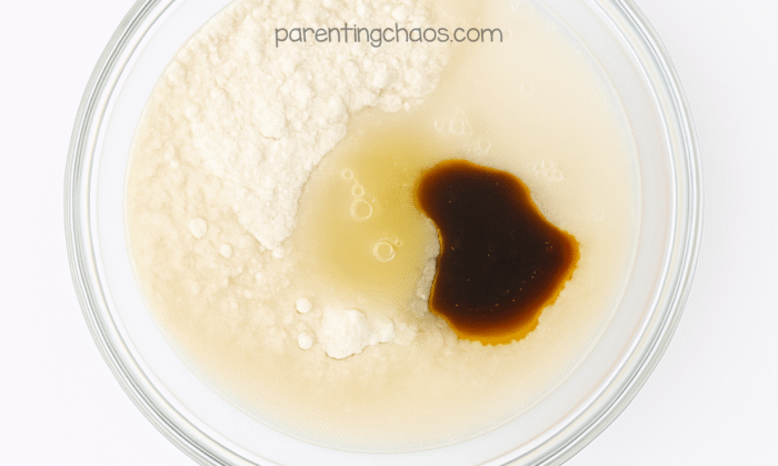 A glass bowl showing sugar cookie playdough ingredients before mixing. Flour, oil, water, and vanilla extract are visible with small bubbles forming on the surface. Text reads “parentingchaos.com.”