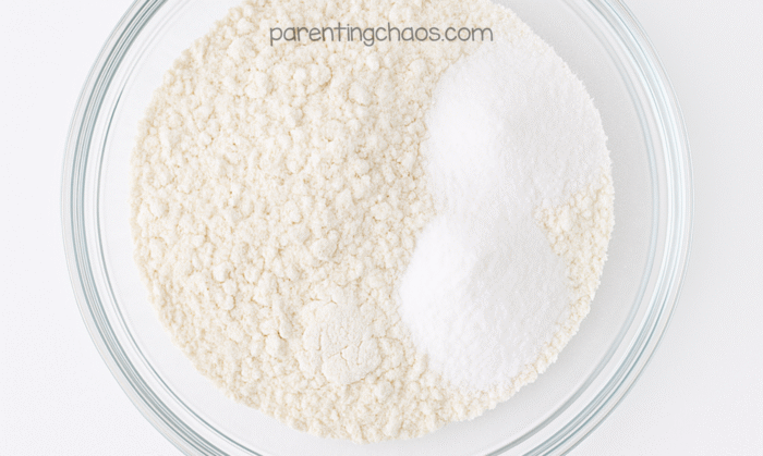 A glass mixing bowl filled with dry ingredients for sugar cookie playdough — flour and salt sitting side by side on a white background. Text reads “parentingchaos.com.”
