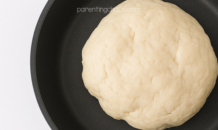 A ball of freshly cooked sugar cookie playdough resting in a dark nonstick pan, soft and smooth in texture. Text reads “parentingchaos.com.”