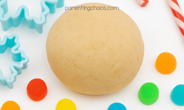 A tan ball of sugar cookie playdough surrounded by blue snowflake cookie cutters, candy canes, and colorful gumdrops. Text reads “parentingchaos.com.”