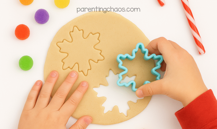 A child’s hands press a blue snowflake cookie cutter into rolled-out sugar cookie playdough, surrounded by gumdrops and candy canes. Text reads “parentingchaos.com.”