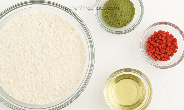 Glass bowls filled with flour, green powder, red heart sprinkles, and oil arranged on a white surface.